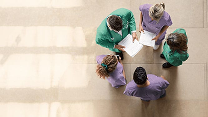 Top view of 5 healthcare professionals in green and purple scrubs standing together and reviewing documents. 